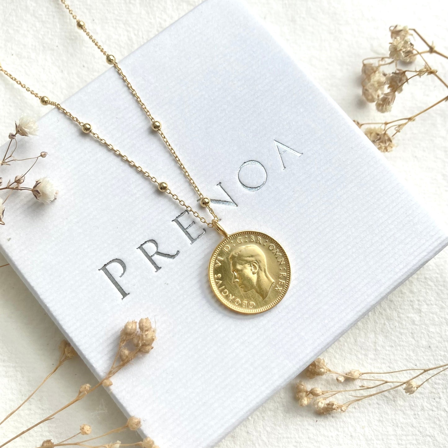 Gold necklace with a pendant on a white card with 'PRENOA' branding, surrounded by dried flowers.