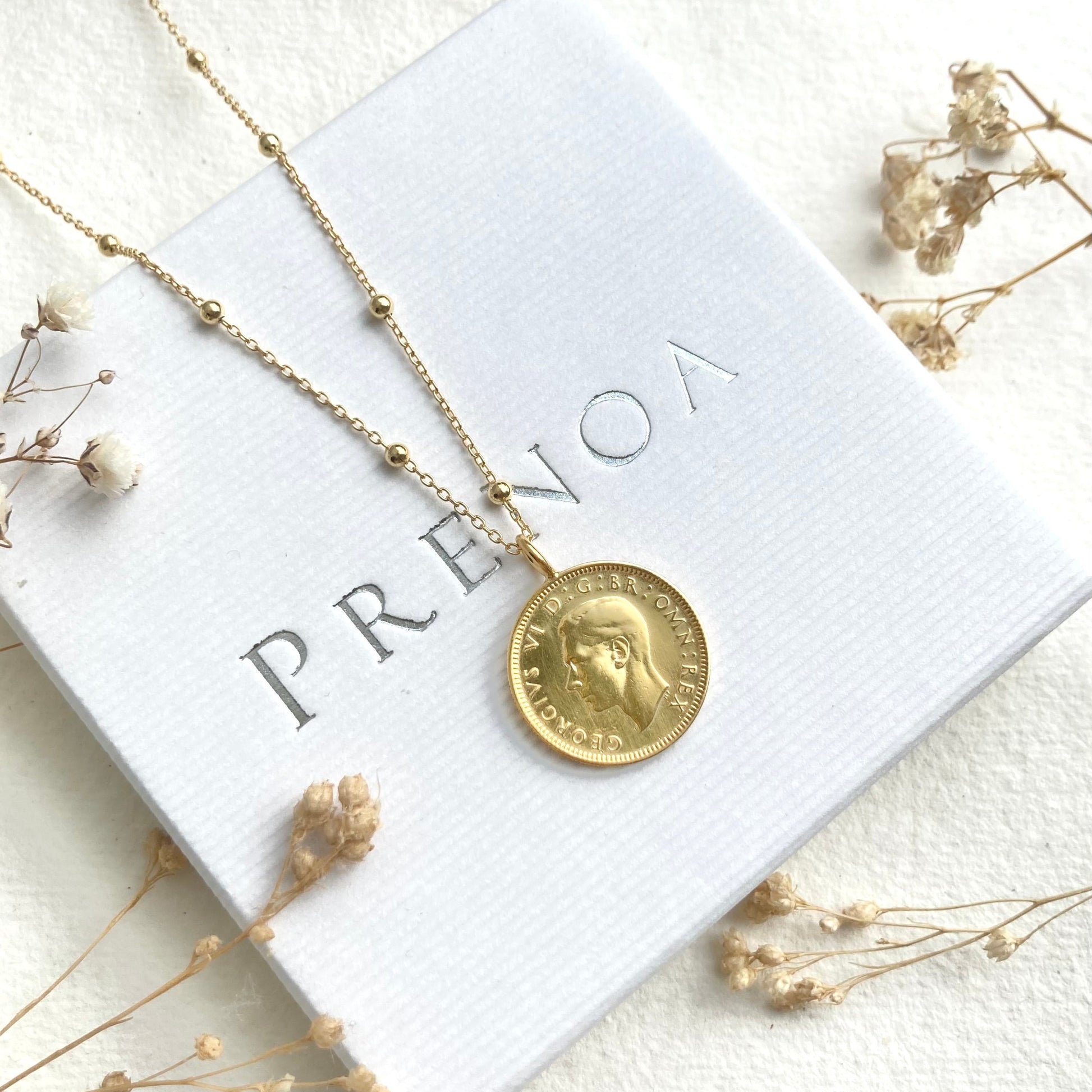 Gold necklace with a pendant on a white card with 'PRENOA' branding, surrounded by dried flowers.