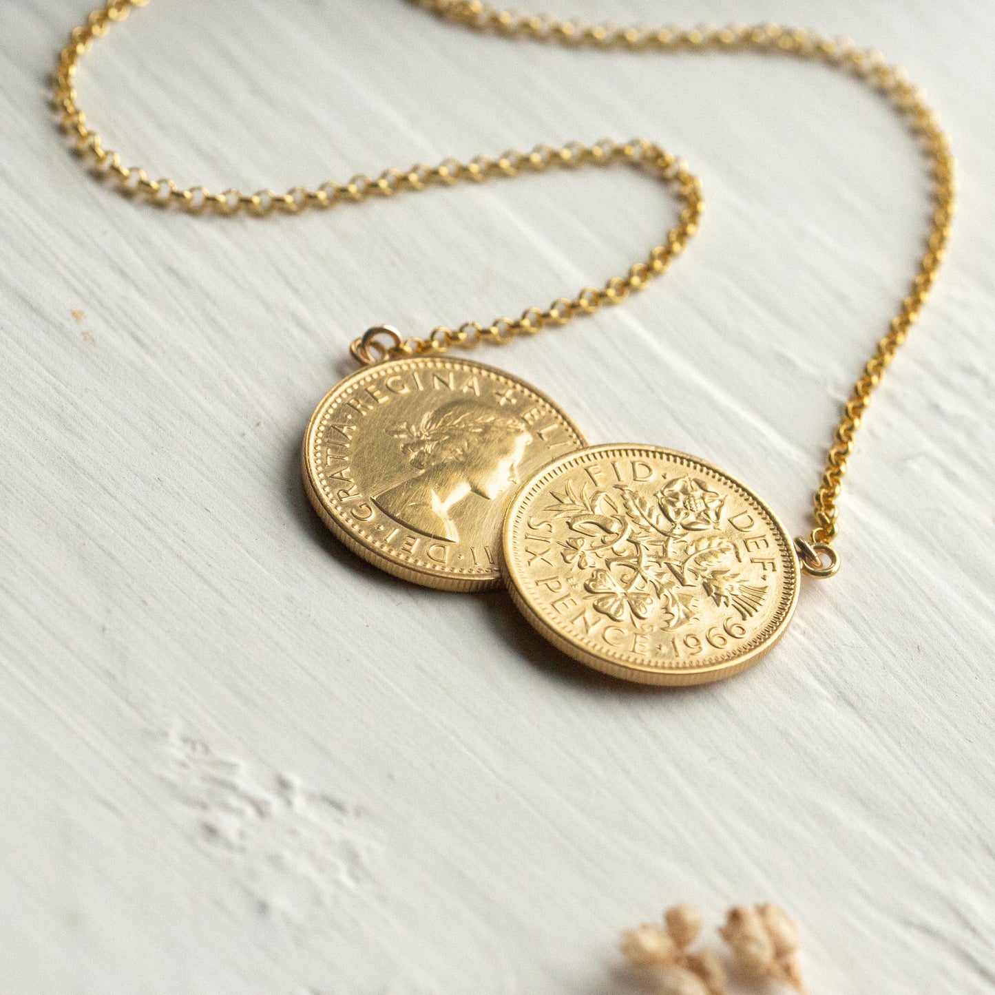 Gold coin necklace on a light wooden surface with dried flowers.