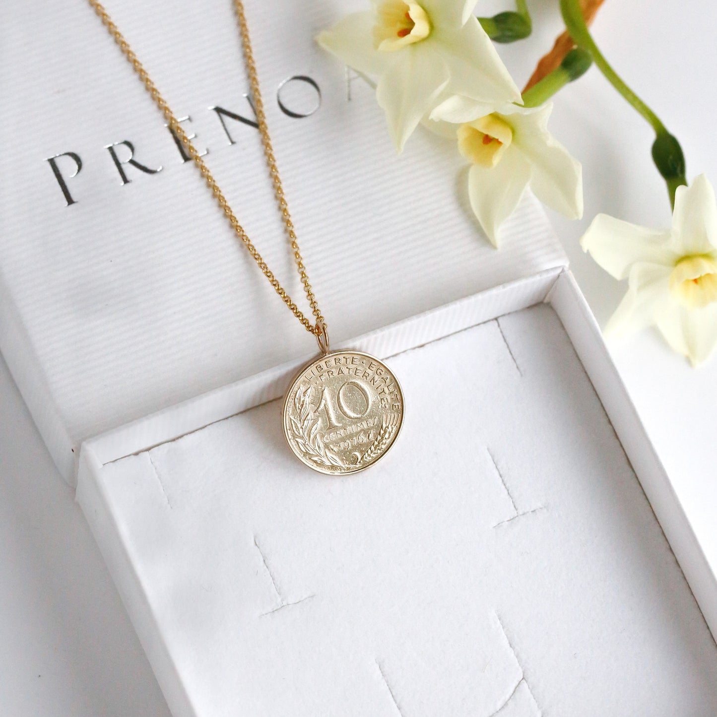 Gold French necklace with a coin pendant on a white box with Prenoa branding, accompanied by flowers.
