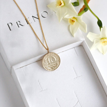 Gold French necklace with a coin pendant on a white box with Prenoa branding, accompanied by flowers.