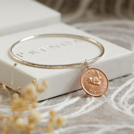 Silver bangle with a bronze farthing coin pendant on a white box with 'Prenoa' branding, on a textured beige surface.