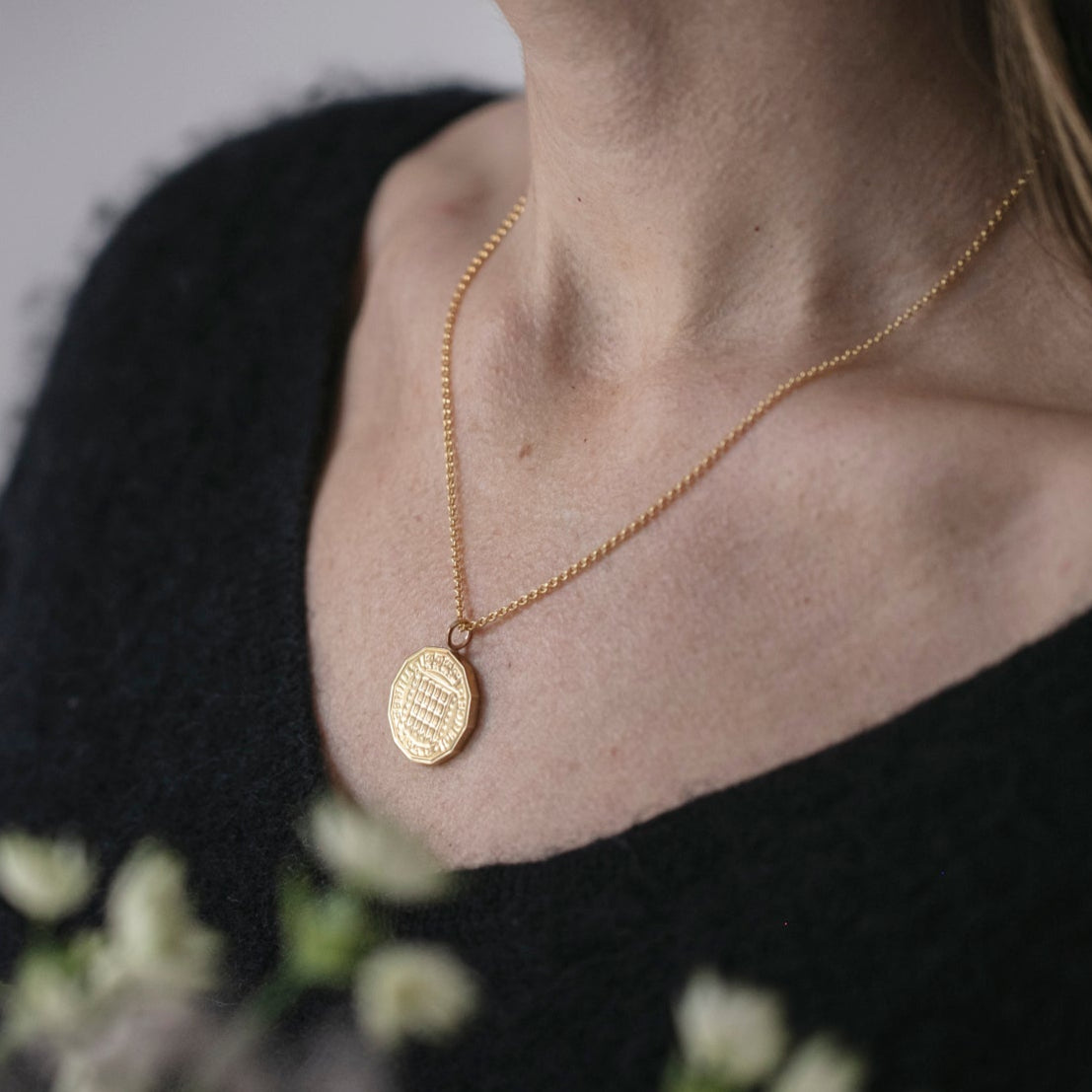 Person wearing a gold necklace with a pendant, standing next to white flowers against a neutral background