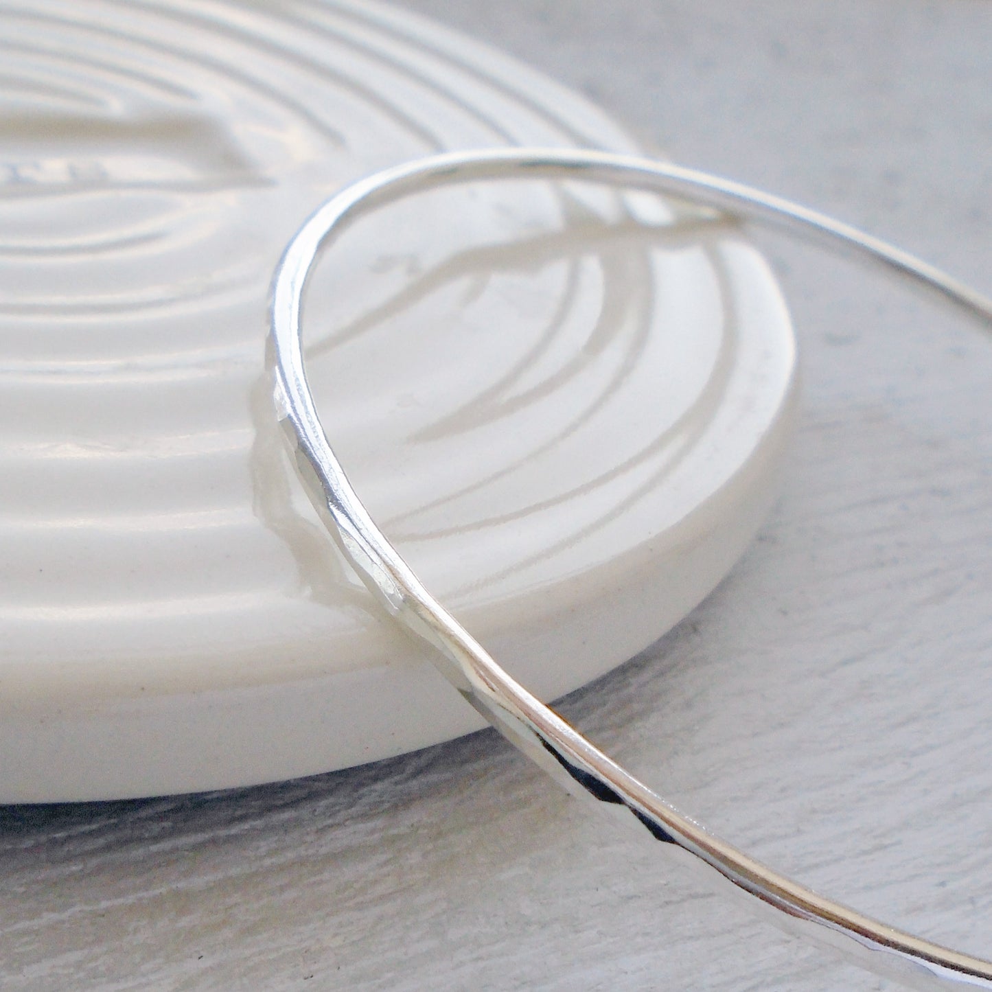 Prenoa's signature lighting hammered coin bangle, ideal for stacking. Showing on a white coaster with light capturing the silver and a wooden painted background.