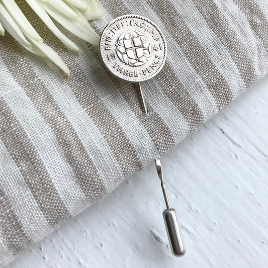 A silver-colored twisted stick pin lapel pin with a safety stopper, featuring the English rose and flag, placed on a grey textured surface with a white flower behind it.