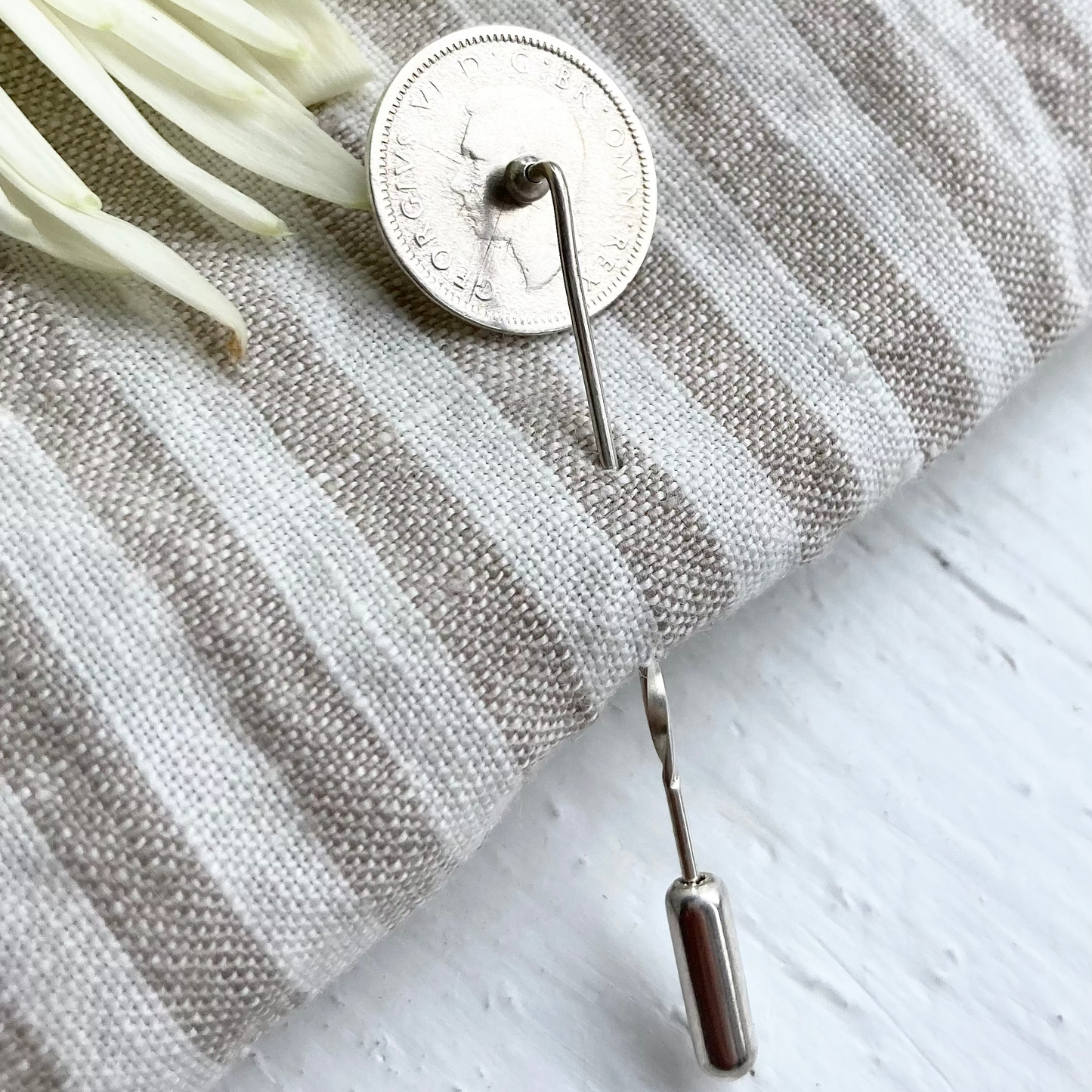 A silver coin lapel pin on a striped linen cloth with white background and suggestion of a flower in the top left.